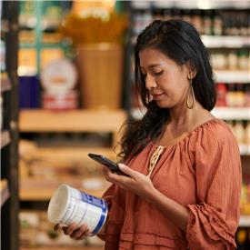At a supermarket, a dark haired woman in orange blouse uses her mobile device to scan a QR code on a blue tin of food At a supermarket, a dark haired woman in orange blouse uses her mobile device to scan a QR code on a blue tin of food